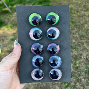 May include: A set of eight glass eye cabochons in various colors, including green, blue, and purple, arranged on a black rectangular backing. Each eye has a reflective, glossy surface with a dark pupil.