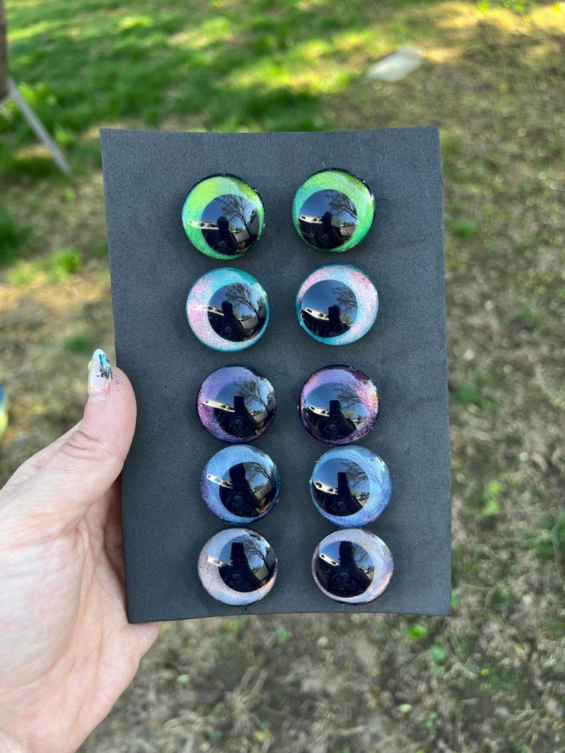 May include: A set of eight glass eye cabochons in various colors, including green, blue, and purple, arranged on a black rectangular backing. Each eye has a reflective, glossy surface with a dark pupil.