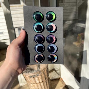 May include: A selection of glass eyes in various colors, including green, blue, and purple, are arranged on a black card. Each eye has a dark pupil and a colorful iris. The eyes are round and appear to be for crafting or doll making.
