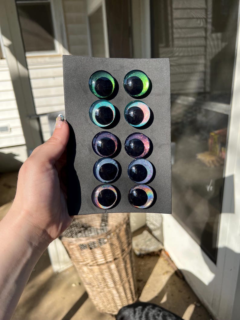 May include: A selection of glass eyes in various colors, including green, blue, and purple, are arranged on a black card. Each eye has a dark pupil and a colorful iris. The eyes are round and appear to be for crafting or doll making.