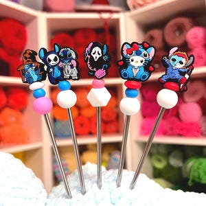 May include: Set of five silver knitting stitch markers with cartoon character charms. Each marker features a different character, including Stitch, Hello Kitty, and a Ghostface design. The markers have colorful bead accents and are displayed on a blue yarn surface.