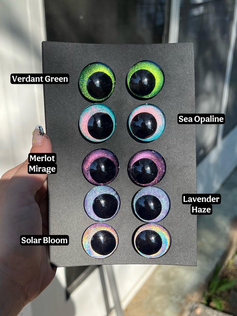 May include: A selection of colorful, reflective doll eyes in various shades, including Verdant Green, Sea Opaline, Merlot Mirage, Lavender Haze, and Solar Bloom. Each eye features a glossy black pupil. The eyes are displayed on a black background.