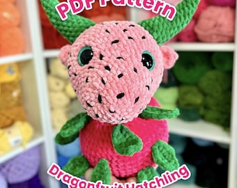 Puede incluir: Un juguete de peluche de dragón de fruta hecho a ganchillo con una cabeza rosa texturizada, motas negras, cuernos verdes y un cuerpo redondo de color rosa oscuro. El texto PDF Pattern y Dragonfruit Hatchling es visible en blanco con borde rosa.