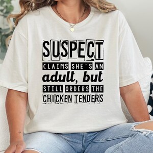 Puede incluir: Camiseta blanca con texto negro que dice "SUSPECT CLAIMS SHE'S AN adult, BUT STILL ORDERS THE CHICKEN TENDERS".