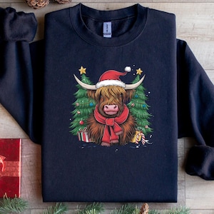 Peut inclure: Un sweat-shirt bleu marine avec un motif de vache des Highlands festive. La vache porte un bonnet de Père Noël et une écharpe rouge, avec des sapins de Noël et des cadeaux en arrière-plan. Un design coloré pour les fêtes.