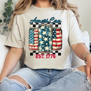 Puede incluir: Camiseta beige con un diseño de tres latas decoradas con motivos de la bandera estadounidense. El texto "Ameri Can" está encima de las latas y "EST. 1776" debajo. La persona lleva vaqueros azules.