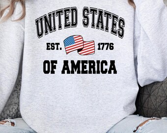 May include: Light gray sweatshirt with the text "UNITED STATES OF AMERICA" in black, arched across the top. Below the text is "EST. 1776" and a waving American flag. The sweatshirt is a crew neck style.