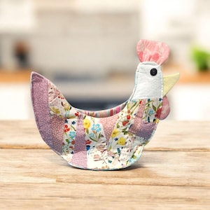 May include: A handmade fabric chicken-shaped basket. The basket features a patchwork design with floral and solid pink fabrics. It has a white head with a black button eye, a yellow beak, and a pink comb and wattle. The basket sits on a wooden surface.