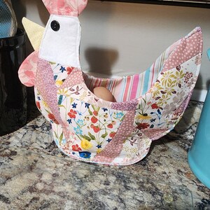 May include: A handmade fabric chicken-shaped egg basket. The basket features a patchwork design with floral and striped patterns in pink, white, and yellow. A single egg rests inside the basket.