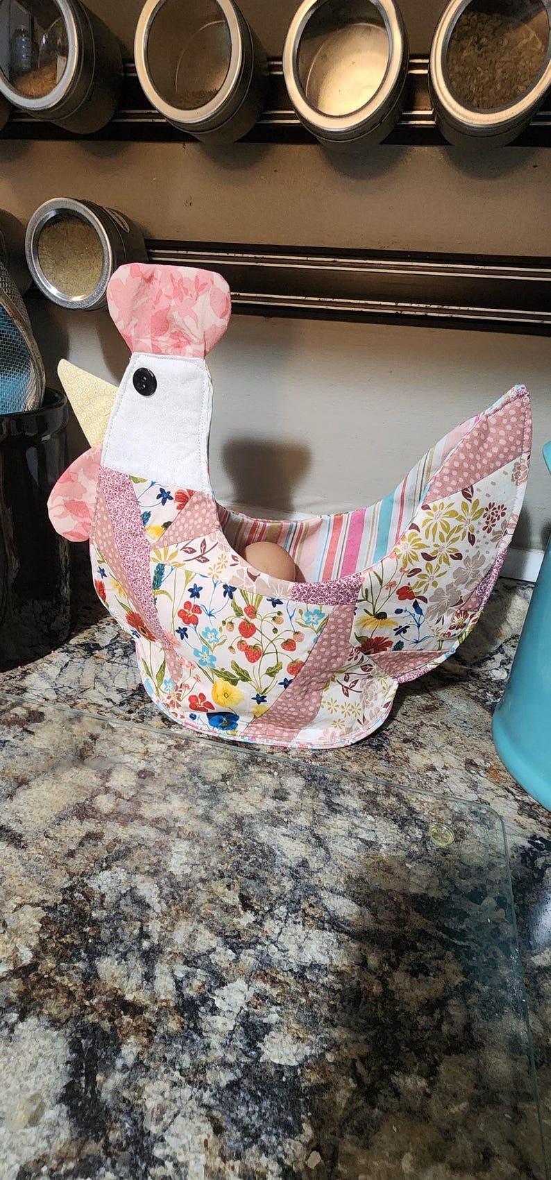 May include: A handmade fabric chicken-shaped egg basket. The basket features a patchwork design with floral and striped patterns in pink, white, and yellow. A single egg rests inside the basket.