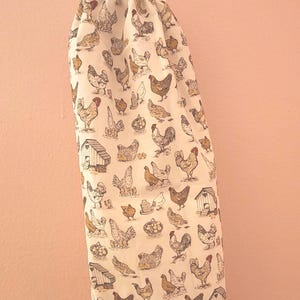 May include: A white drawstring bag with a repeating pattern of chickens, hen houses, and eggs in shades of brown and gray. The bag is hanging from a white hook against a pink wall. The bag is made of fabric.