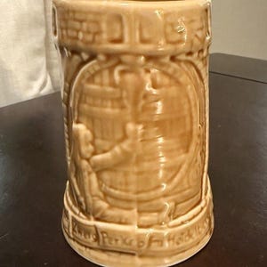 May include: Tan ceramic beer stein with a detailed relief design. The stein features a barrel scene with figures and text around the base. The cylindrical mug has a slightly flared base and a textured surface.