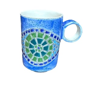 May include: Blue ceramic mug with a mosaic design in shades of blue and green. The mug has a round handle and a white interior. The mosaic pattern is centered on the mug's body.