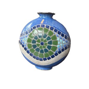 May include: A round, blue ceramic vase with a mosaic design. The mosaic features a central green and blue circular pattern, surrounded by blue and green squares. The vase has a textured, white band near the base and a dark rim.