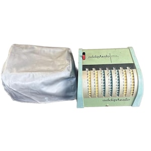 May include: A vintage light green Addipresto adding machine with vertical number wheels and a red switch, accompanied by a matching light gray fabric dust cover. The device features the brand name printed in black on the front metal casing.