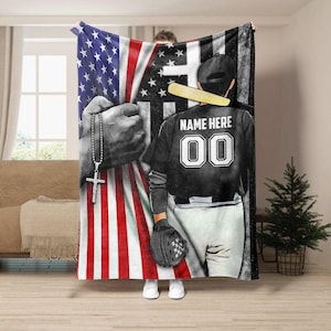 May include: A baseball-themed blanket with an American flag design and a baseball player graphic. The player's jersey has the text "NAME HERE" and the number "00". The blanket also includes a black and white image of hands holding a rosary.