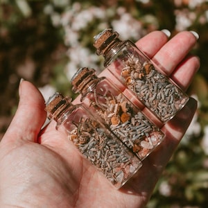 May include: Three small glass bottles filled with dried herbs and flowers, each sealed with a cork. The bottles are held in a hand, with a blurred green and white background. The contents of the bottles appear to be a mix of lavender, chamomile, and other botanicals.