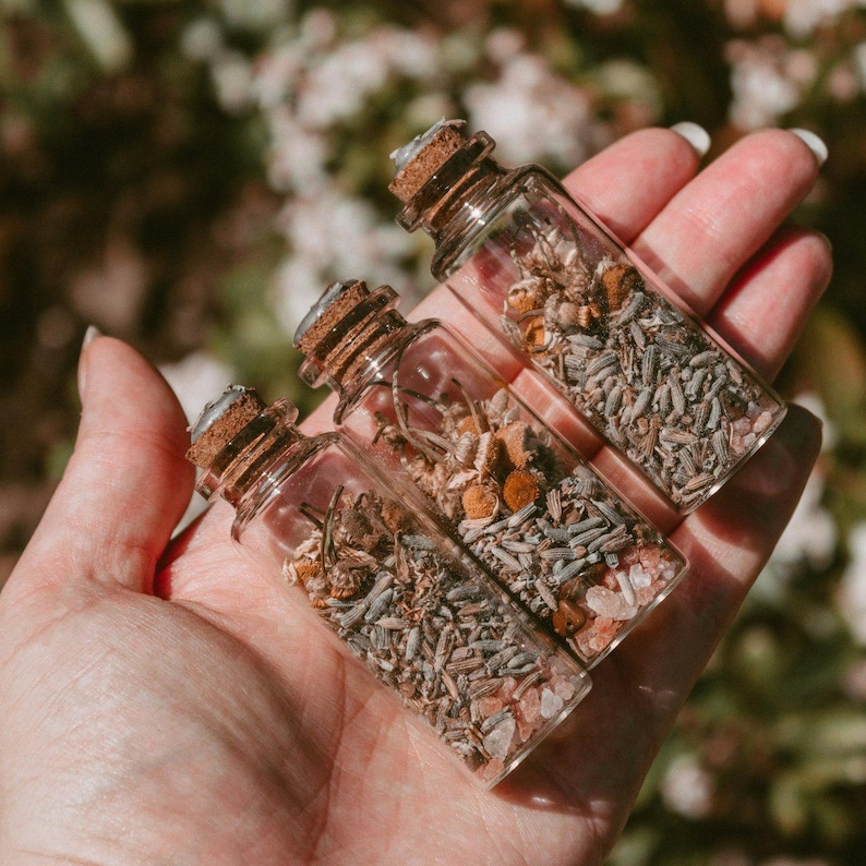 May include: Three small glass bottles filled with dried herbs and flowers, each sealed with a cork. The bottles are held in a hand, with a blurred green and white background. The contents of the bottles appear to be a mix of lavender, chamomile, and other botanicals.