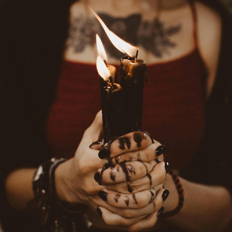 May include: Two black candles with flames burning brightly are held in hands with black nail polish. The wax is dripping down the candles and onto the hands. The background is a dark red.