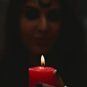 May include: A lit red candle with a bright yellow flame. The candle is held in front of a dark background, with a blurred figure in the background. The candle's wax is a deep red color.