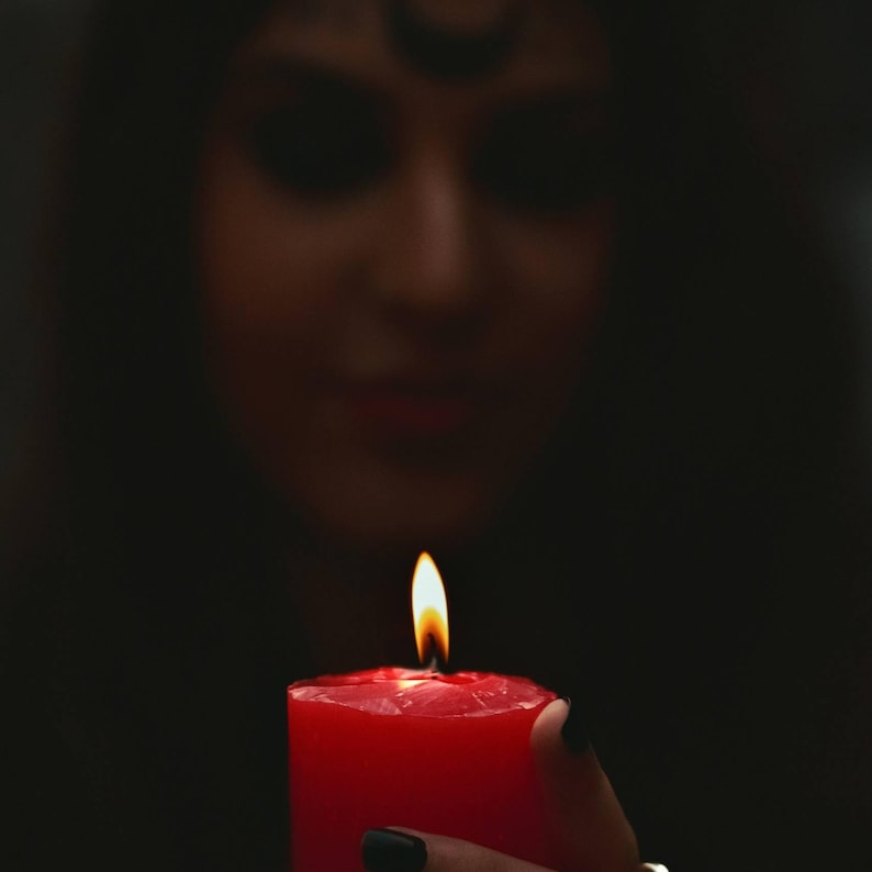 May include: A lit red candle with a bright yellow flame. The candle is held in front of a dark background, with a blurred figure in the background. The candle's wax is a deep red color.