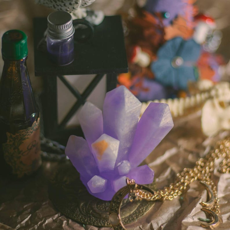 May include: A cluster of purple crystal formations with a gold-colored pendant necklace. A small bottle with a green cap and a black lantern are also visible. The items are arranged on a textured, brown surface.