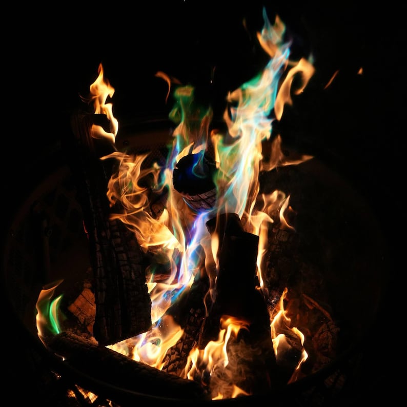 May include: A close-up shot of a campfire with flames in shades of orange, yellow, green, and blue. The fire is burning in a metal fire pit, with logs of wood visible. The background is dark.