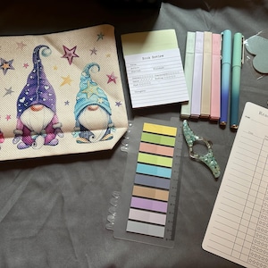May include: A collection of stationery items, including a beige pencil case with gnome illustrations, sticky notes, highlighters, pens, a heart-shaped bookmark, and a reading log. The items are arranged on a grey surface.