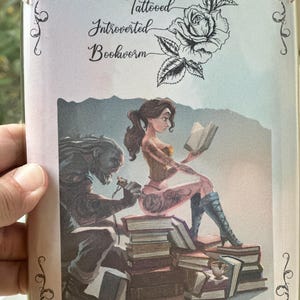 Puede incluir: Una ilustración colorida de una mujer que se tatúa la pierna por un hombre. La mujer está sentada en una pila de libros y sostiene un libro en su mano. El texto "Tattooed Introverted Bookworm" está en la parte superior de la imagen.
