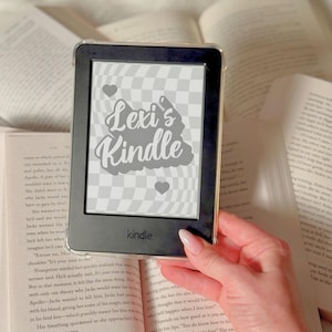 May include: A black and white Kindle e-reader with a custom design that says "Lexi's Kindle" in a gray cloud shape with two small hearts. The Kindle is sitting on top of a book.
