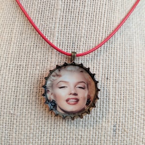 May include: A red cord necklace with a gold-toned bottle cap pendant featuring a black and white image of Marilyn Monroe.