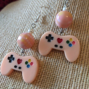 May include: A pair of pink and white dangle earrings featuring a pink and white swirl bead and a pink plastic video game controller with black buttons and a red heart.