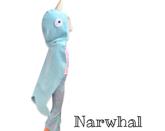 May include: A child wearing a light blue narwhal costume with a white horn and a large white eye. The costume is a hooded cape with a long tail. The child is wearing a pair of iridescent leggings. The text "Narwhal" is printed below the child.