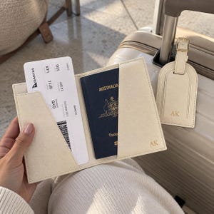 May include: A cream-colored passport holder and matching luggage tag, both featuring the gold-embossed initials AK. The holder contains a dark blue Australian passport and a Qantas boarding pass for a flight from QYD to LAX.