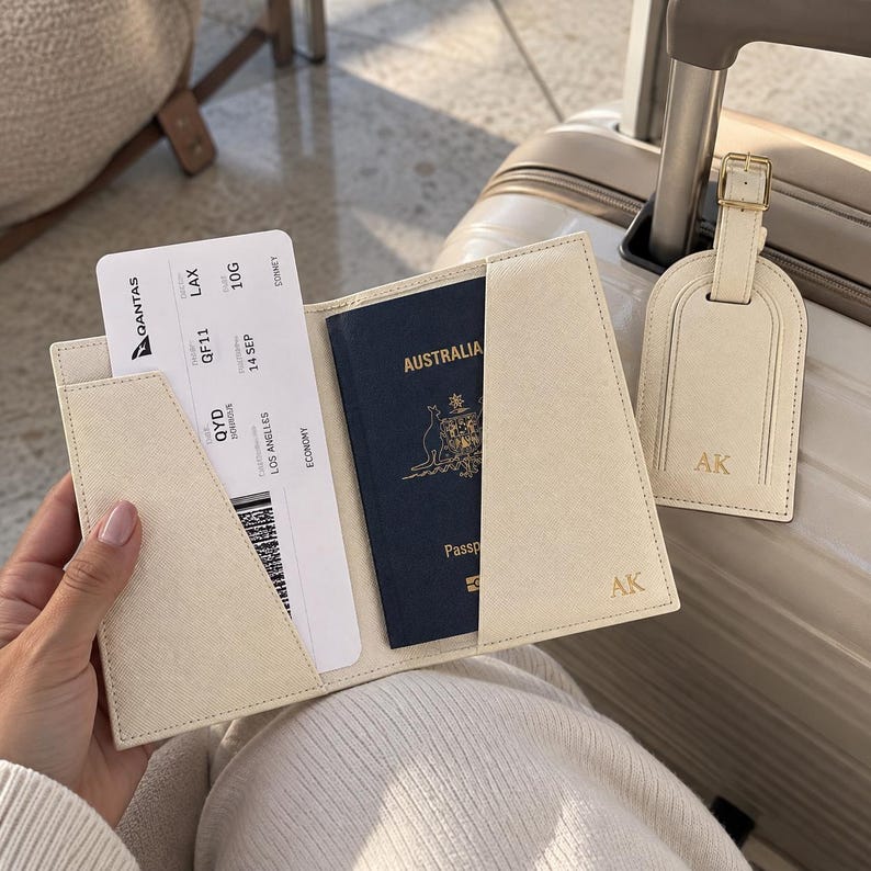 May include: A cream-colored passport holder and matching luggage tag, both featuring the gold-embossed initials AK. The holder contains a dark blue Australian passport and a Qantas boarding pass for a flight from QYD to LAX.