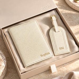 May include: A cream-colored passport holder and luggage tag set with a textured finish, presented in a matching gift box. The passport holder features the name Sophia in gold, while the luggage tag displays the initials SM in gold.