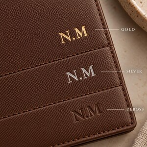 May include: A dark brown textured leather surface featuring three horizontal rows of the monogram N.M. The top row is in gold foil, the middle row is in silver foil, and the bottom row is a plain debossed impression. Each row includes a label.