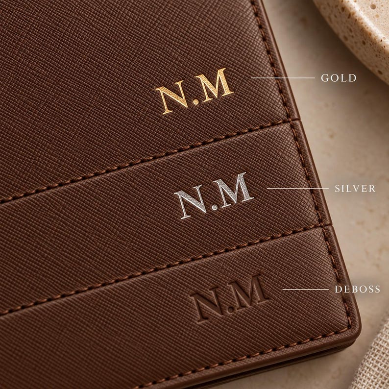 May include: A dark brown textured leather surface featuring three horizontal rows of the monogram N.M. The top row is in gold foil, the middle row is in silver foil, and the bottom row is a plain debossed impression. Each row includes a label.