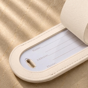 May include: A cream-colored luggage tag with a textured surface and a rounded end. The tag features a white insert with printed lines and the labels Name, Address, and Phone. A matching strap is attached to the top of the luggage tag.