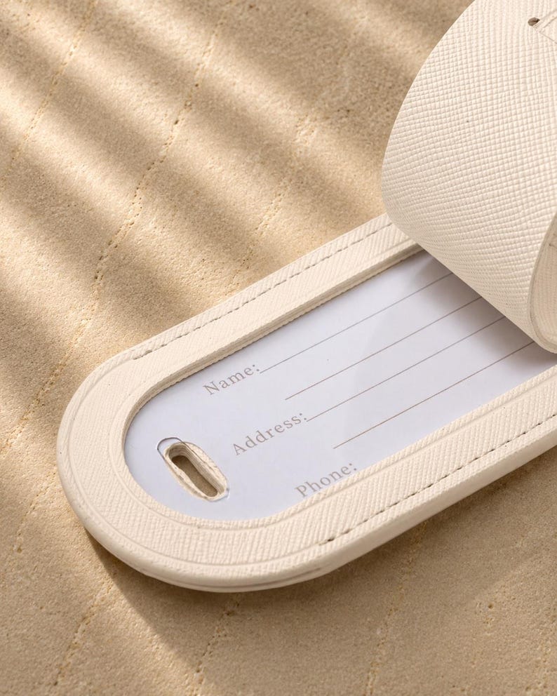 May include: A cream-colored luggage tag with a textured surface and a rounded end. The tag features a white insert with printed lines and the labels Name, Address, and Phone. A matching strap is attached to the top of the luggage tag.