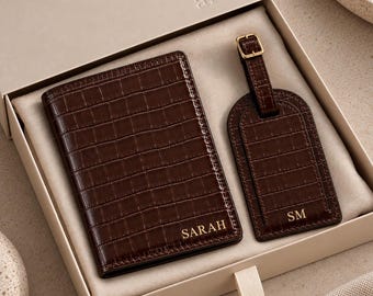 May include: A dark brown crocodile-embossed passport holder and luggage tag set presented in a gift box. The passport holder features the name SARAH in gold lettering, while the luggage tag displays the initials SM in gold, both with gold hardware.