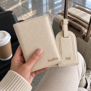 May include: A cream-colored passport holder with the name Sophia embossed in gold and a matching luggage tag with the initials SM embossed in gold. Both items feature a textured finish and are made of a durable, synthetic material.