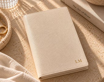 May include: A cream-colored, textured leather notebook featuring the gold-embossed initials "LM" in the bottom right corner. The rectangular journal has a clean, minimalist cover design with a visible grain pattern and stitched edges.