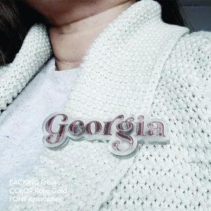 May include: A personalized nameplate with the word "Georgia" in a rose gold color with a frosted backing. The nameplate is made with a decorative script font.