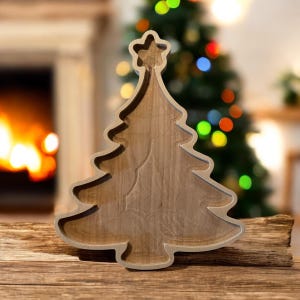 May include: Wooden Christmas tree-shaped tray with a star on top. The tray is made of light brown wood and has a carved design. It is placed on a wooden surface with a blurred background of a fireplace and a Christmas tree.