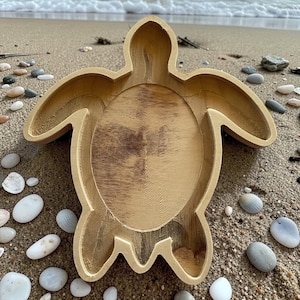 May include: A wooden turtle-shaped tray with a natural wood grain finish. The tray has a shallow, bowl-like interior and is surrounded by the turtle's shell and flippers. The tray is resting on a sandy surface with small pebbles and seashells scattered around it.