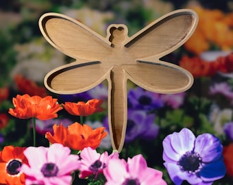 May include: A wooden dragonfly-shaped decorative item with a light brown finish. The dragonfly is set against a backdrop of colorful flowers, including red, pink, and purple anemones. The wings and body are clearly defined.