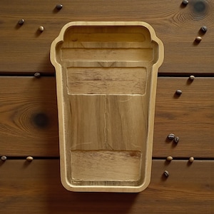 May include: A wooden tray in the shape of a coffee cup, with a carved design. The tray is light brown and sits on a wooden surface. Coffee beans are scattered around the tray.