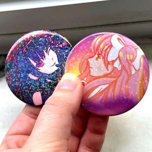 May include: Two round, colorful buttons with anime-style illustrations. One button features a character with dark hair and a white face, set against a sparkly blue background. The other has a pink and orange gradient with a pink-haired character.