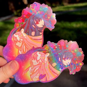 以下が含まれることがあります： ピンク、パープル、ブルーのグリッター背景にアニメキャラクターが描かれたホログラフィックステッカー2枚。キャラクターは星とハートで囲まれています。